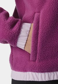 Persona che indossa una giacca in pile viola con una mano parzialmente infilata nella tasca anteriore, che mostra una manica interna bianca.