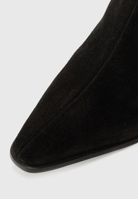 Bottine en suède noir avec un bout pointu, texture lisse et semelle noire fine, mettant en valeur des lignes de design épurées et minimalistes.