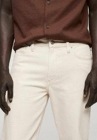 Pantaloni in denim color crema con vestibilità dritta, design a cinque tasche e cuciture rinforzate. Abbinati a una camicia marrone a maniche lunghe con bottoni.