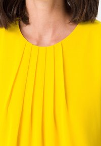 Blusa gialla con pieghe verticali sul colletto. Tessuto morbido con una superficie liscia, caratterizzata da un colletto rotondo e senza elementi hardware visibili.