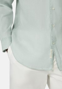 Camicia button-up verde menta realizzata in tessuto texturizzato, con orlo curvo e una piccola etichetta sul lato. Abbinata a pantaloni bianchi.