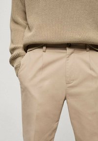 Beige, op maat gemaakte broek van gladde stof, met voorplooien en zijzakken. Daarboven is een gebreide beige trui.