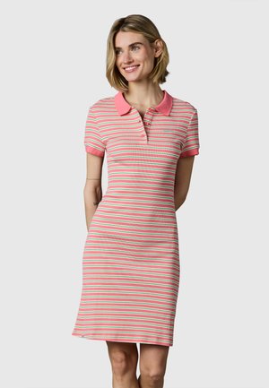 Lachende vrouw die een roze, witte en groene gestreepte polojurk met korte mouwen draagt, staand tegen een effen achtergrond.