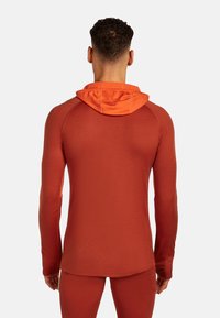 Hombre con una sudadera ajustada de manga larga en tonos naranja quemado, con la capucha baja, visto desde atrás sobre un fondo blanco liso.