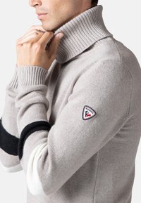 Maglione a collo alto in maglia beige chiaro con texture a coste. Presenta una striscia nera e bianca e una patch con logo sulla parte superiore del braccio.