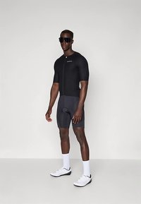 Combinaison de cyclisme noire à manches courtes avec zip, associée à un short gris foncé. Des chaussures de sport blanches et des lunettes de soleil complètent la tenue.