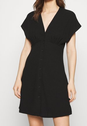 Kvinde iført en kort sort knap-ned kjole med V-hals og korte ærmer, hænder afslappet ved siderne mod en ensfarvet baggrund.