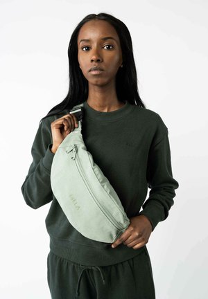 Vrouw in donkergroene trui en broek houdt lichtgroene MELA schoudertas vast tegen een witte achtergrond.