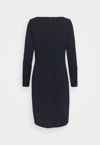 Robe noire à manches longues longueur genou avec fermeture éclair dans le dos et silhouette ajustée sur un fond clair uni.
