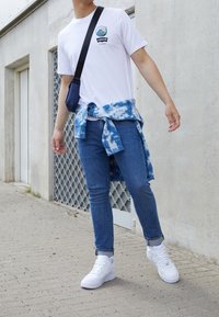 Bílá tričko s grafickým motivem vlny, modrá kostičkovaná košile zavázaná kolem pasu, modré džíny a bílé tenisky. Doplněk tvoří malá černá taška.