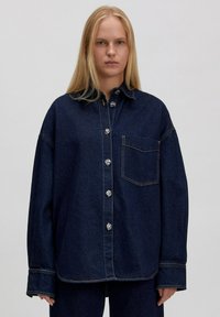 Donkerblauwe denim overhemd met een kraag, een voorzak en grote knopen. Losse pasvorm met zichtbare stiksels en zijsplitten aan de zoom.