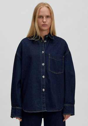 Donkerblauwe denim overhemd met een kraag, een voorzak en grote knopen. Losse pasvorm met zichtbare stiksels en zijsplitten aan de zoom.