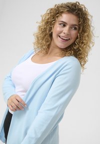 Hellblauer, langärmliger Cardigan mit offenem Vorderteil, aus weichem Material, über einem weißen Top getragen. Strukturiert, mit entspannter Passform.
