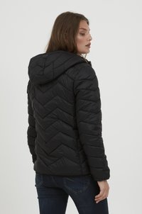 Sort puffjakke med hætte, chevron-quiltet mønster og elastiske manchetter. Båret med mørke jeans, set fra ryggen.