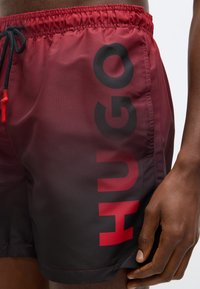 Κόκκινα και μαύρα μαγιό με σχέδιο βαθμίδωσης, με μεγάλο μαύρο λογότυπο "HUGO" στο πλάι, ελαστική μέση με κορδόνι, λεία υφή.