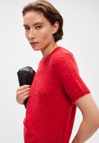 Piros, rövid ujjú pulóver, puha, bolyhos anyagból készült, kerek nyakkivágással. A modellen egy fekete, texturált részletekkel rendelkező clutch táska van.