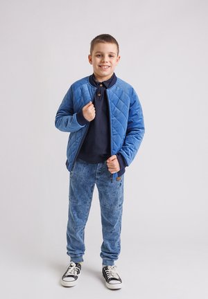 Uśmiechnięty chłopiec w niebieskiej pikowanej kurtce na granatowej koszulce, niebieskich dżinsach i czarno-białych trampkach, stojący na tle jednolitego jasnego tła.