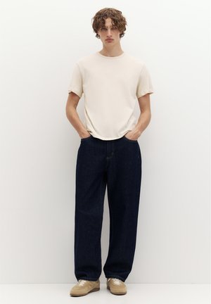 Jeune homme aux cheveux bouclés portant un t-shirt beige uni, un jean bleu foncé ample et des baskets beiges, debout les mains dans les poches.