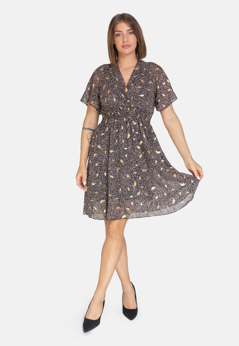 Vestido negro con mangas cortas y vuelos, patrón floral en amarillo y blanco, cintura ceñida, falda fluida, combinado con tacones negros de punta.