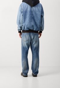 Denim jacket in light blue with a black hooded sweatshirt underneath. Blue jeans with a relaxed fit, displayed from the back. Black shoes.