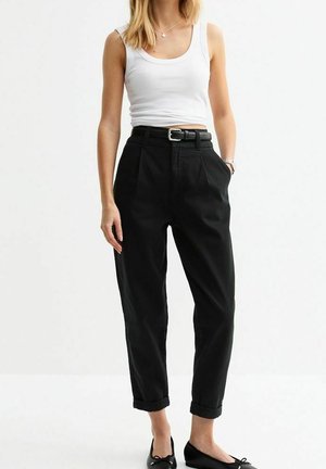 Femme portant un haut blanc sans manches rentré dans un pantalon noir taille haute, fuselé, avec une ceinture, des ourlets retroussés et des ballerines noires.