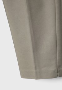 Gros plan sur une jambe de pantalon en tissu beige avec pli visible et détail de couture sur fond uni.