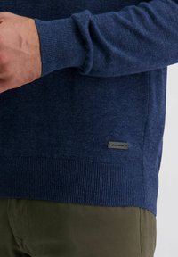 Dunkelblaues Strickpullover mit gerippten Bündchen und Saum, mit einem kleinen schwarzen Etikett an der Seite. Weiche Textur, taillierte Passform.