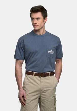 Uomo che indossa una maglietta blu Sebago Docksides infilata in pantaloni cargo beige con cintura marrone, che guarda leggermente di lato.