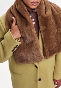 Person iført en olivengrøn frakke med et stort, brunt kunstlæder-halstørklæde og en mauve strikket højhalset trøje, som holder halstørklædet nær kraven.