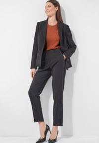 Frau in schwarzem, tailliertem Blazer und Hose mit rustfarbenem Oberteil und schwarzen High Heels, Stehend vor weißem Hintergrund.