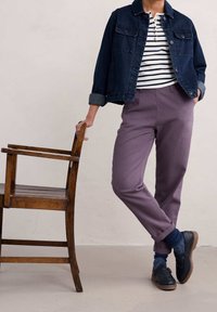 Jeansjacka i mörkblått, randig vit och svart skjorta, lila byxor, blå strumpor, och svarta skor med bruna sulor, stående bredvid en trästol.