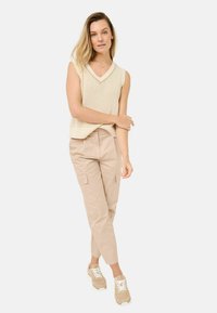 Beige mouwloos gebreid topje met een V-hals, gecombineerd met lichtbeige cargobroeken met zijzakken en crèmekleurige sneakers.