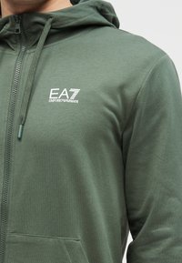 Groene capuchontrui met rits, gemaakt van zachte stof. Voorzien van een capuchon met trekkoord, steekzakken aan de voorkant en een wit logo op de borst. Soepele textuur.