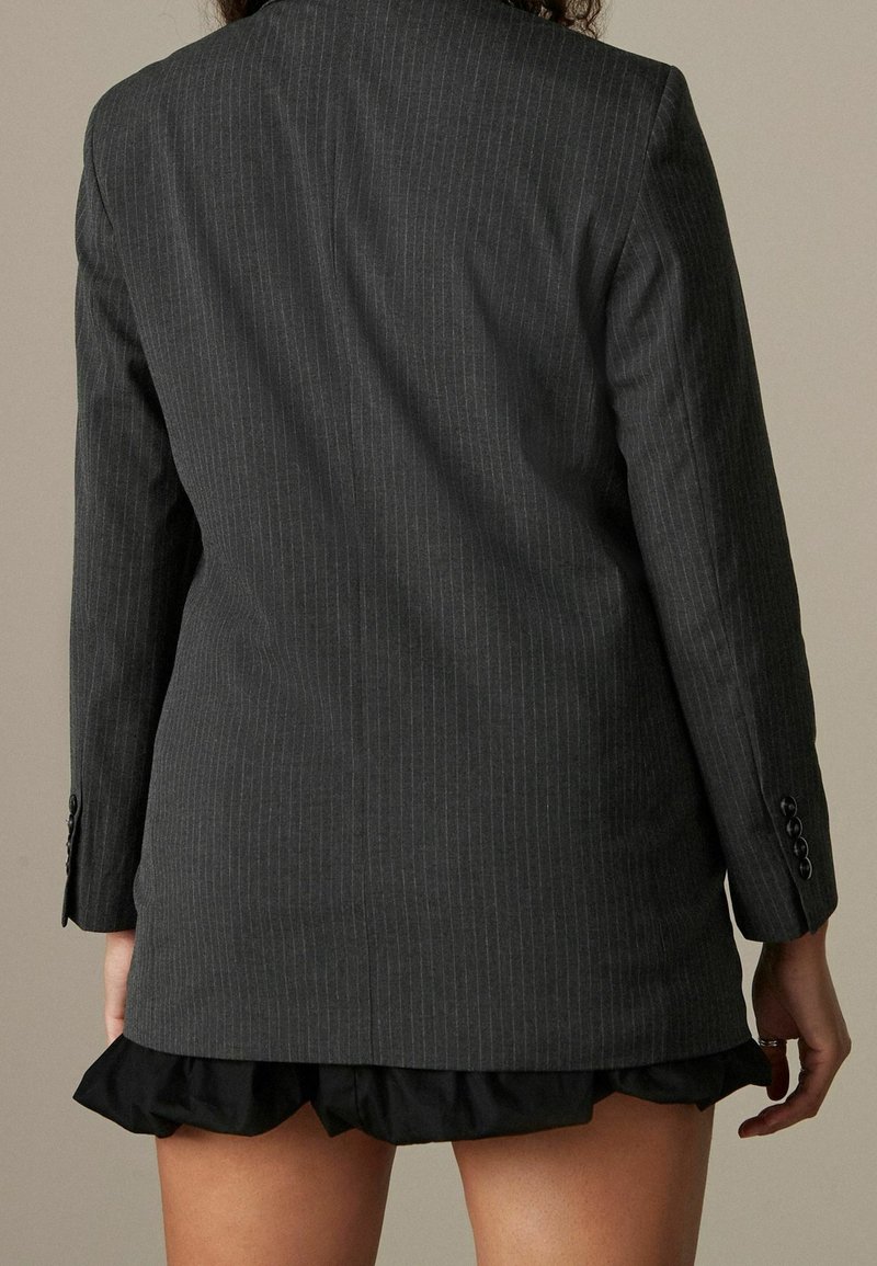 Personne portant un blazer gris foncé à fines rayures sur une jupe noire à volants, vue de dos sur un fond uni.