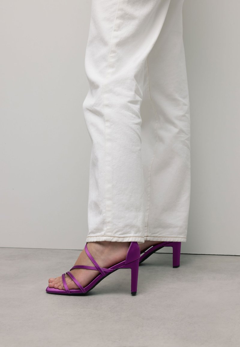 Paarse sandalen met hoge hakken en een strappy ontwerp. De schoen heeft een spitse neus en een slanke stilettohak, gecombineerd met witte jeans.