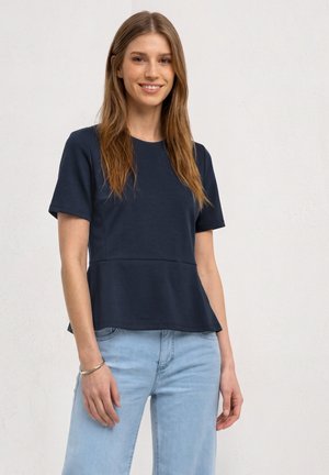 Junge Frau mit langen braunen Haaren, die ein dunkelblaues kurzärmeliges Oberteil und hellblaue Jeans trägt, lächelt vor einem schlichten Hintergrund.