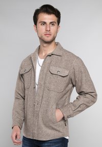 Bruine herringbone jas met twee borstzakken, metalen drukknoopknopen en lange mouwen. Draagt over een wit T-shirt, gecombineerd met blauwe jeans.