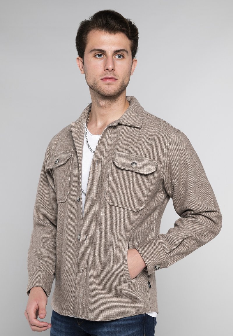 Bruine herringbone jas met twee borstzakken, metalen drukknoopknopen en lange mouwen. Draagt over een wit T-shirt, gecombineerd met blauwe jeans.