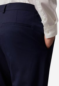 Pantaloni sartoriali blu navy con una texture liscia, dotati di una tasca posteriore e una mano posizionata nella cucitura laterale. Camicia bianca con polsini con bottone.