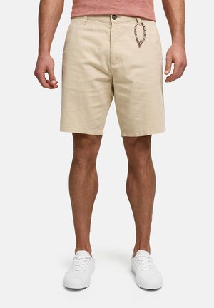 Mand iført beige knælange shorts, hvide sneakers og en laksefarvet skjorte mod en ensfarvet lys baggrund.