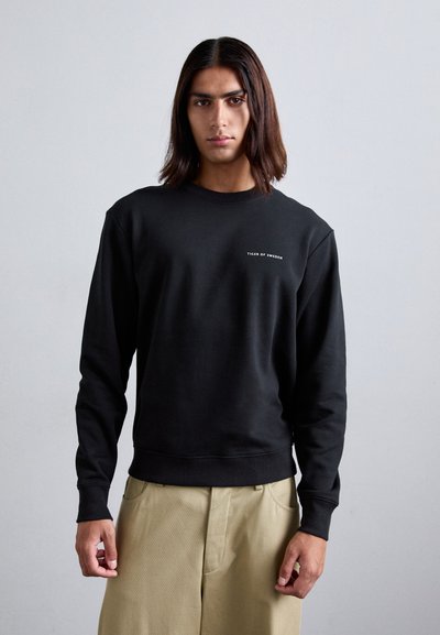 Felpa nera in tessuto morbido, con scollo rotondo e maniche lunghe. Piccolo testo del logo bianco sul petto. Abbinata a pantaloni beige.