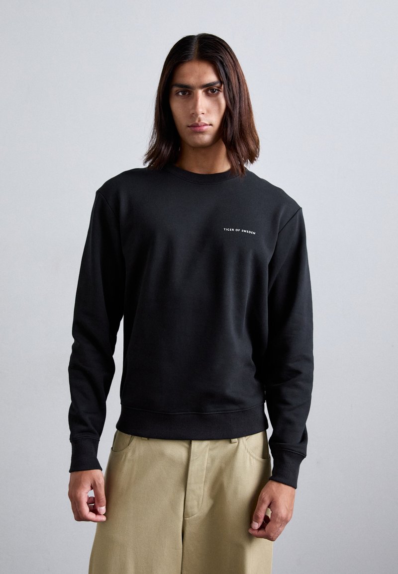 Black sweatshirt in soft fabric, featuring a crew neckline and long sleeves. Small white logo text on the chest. Styled with beige pants.