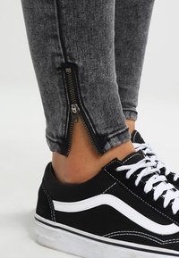 Leggings de mezclilla gris con cremalleras en los tobillos, combinados con zapatillas negras con acentos y cordones blancos. Vista de cerca de la parte inferior de la pierna.