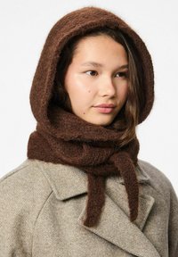 Hnědý, strukturovaný pletený šátek s kapucí a dlouhými vázacími šňůrkami, nošený přes šedý kabát. Materiál se zdá být nadýchaný a teplý, což zvýrazňuje útulný vzhled.