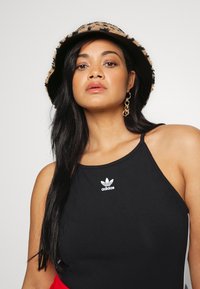 Canotta nera con spalline sottili, con logo adidas bianco. Il modello indossa un cappello a secchiello con stampa leopardata e orecchini a cerchio dorati.