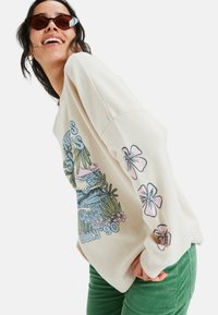 Kermanvärinen huppari, jossa on teksturoitu pinta. Hupparissa on trooppinen graafinen kuvasto sinisen sävyissä ja pinkkejä kukkakoristeita hihoissa.