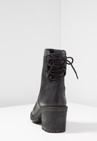Stivale alla caviglia in pelle nera con tacco a blocco, cuciture decorative, lacci sul retro legati a fiocco e zip laterale su una superficie bianca.