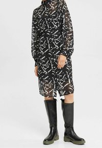 Schwarzes, durchsichtiges Kleid mit abstraktem weißen Muster, langen Ärmeln, hohem Kragen, kombiniert mit knielangen schwarzen und grünen Gummistiefeln.