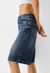 Bermuda in denim blu medio con taglio dritto, orlo sfilacciato e design a cinque tasche. Presenta passanti per cintura e chiusura con bottone.