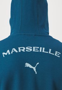 Mėlynas sukapotas su kapišonu, su žodžiu "MARSEILLE" balta spalva ir balta Puma logotipu nugaroje. Su raukšlėta tekstūra ir siuvimo detalėmis.